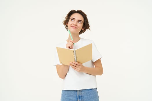 Portrait of creative girl, student doing homework, writing in notebook, standing with her diary, posing over white background.