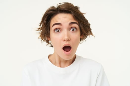 Portrait of woman in shock, drops jaw and stares at camera with mouth wide open, amazed by something, genuine surprise, stands over white background.