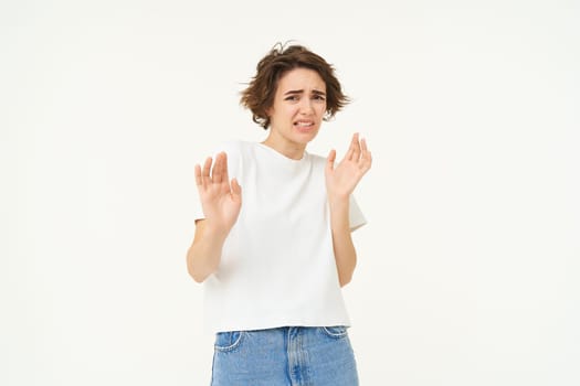 Young reluctant woman steps back, raises hands to refuse, rejects something disgusting, reaction of aversion and dislike, isolated over white background.