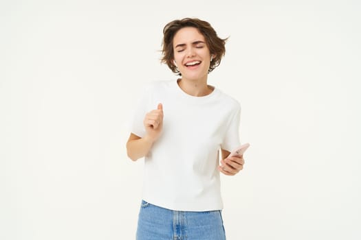 Portrait of woman singing and listening music in wireless headphones, holding mobile phone, having fun, posing over white studio background.