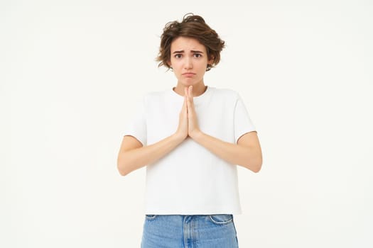 Portrait of girl looking with begging face, holding hands in pray, pleading, making wish, standing over white studio background.