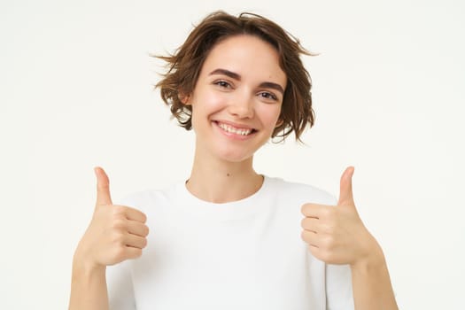 Close up of confident, smiling candid girl, shows thumbs up, recommends, gives positive feedback, approves smth good, stands over white studio background.