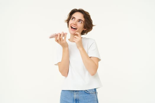 Portrait of brunette woman recording voice message, girl talking on mobile phone, using speakerphone, translating her words, standing over white background.