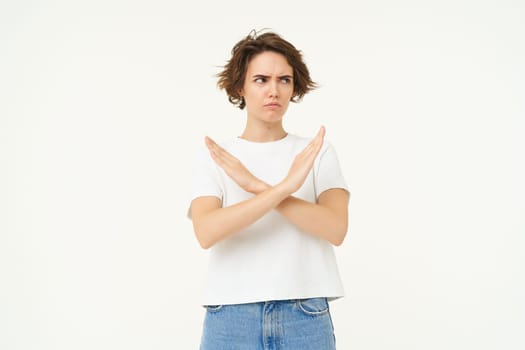 Image of woman expresses dislike, shows cross, stop gesture, disapprove something, prohibit, stands over white studio background.