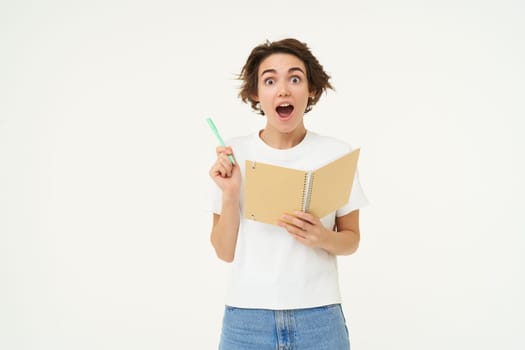 Excited young woman writing down in planner, thinking of solution, says eureka, suggests plan, stands over white studio background. Copy space