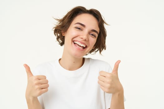 Close up of happy brunette woman, shows thumbs up, approves something, recommends, gives positive feedback, stands over white background.