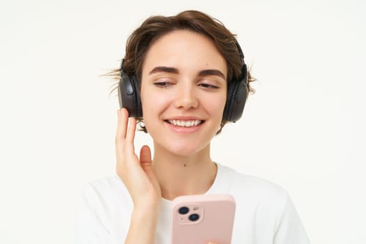 Portrait of happy, smiling beautiful woman, listening to music in wireless headphones, looking at smartphone, choosing song on streaming app, holding mobile phone, standing over white background.