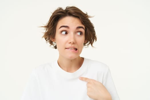 Awkward girl points at herself with guilty look, oops face, stands over white background.