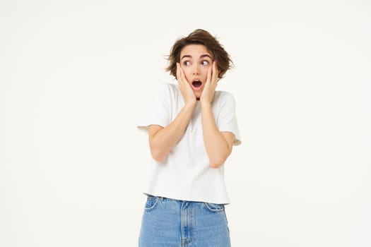 Portrait of brunette woman with shocked, amazed face, drops jaw and gasps worried, looking at camera, isolated over white background.