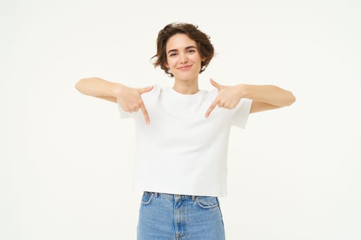 Portrait of cute, happy young woman pointing fingers down, showing advertisement, demonstrating banner on the bottom, standing against white background.