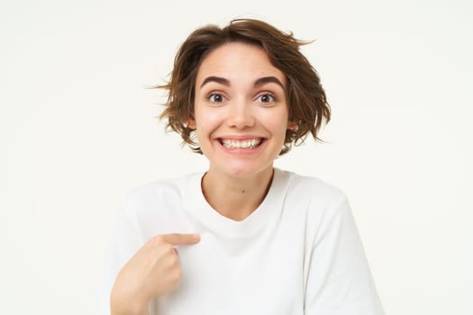 Portrait of girl with surprised face, points at herself amazed, stands isolated over white studio background. Copy space