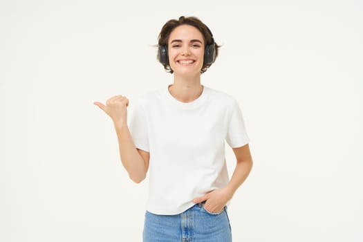 Image of young modern woman, wearing white t-shirt, pointing left, smiling and looking happy, showing advertisement, standing over white background.