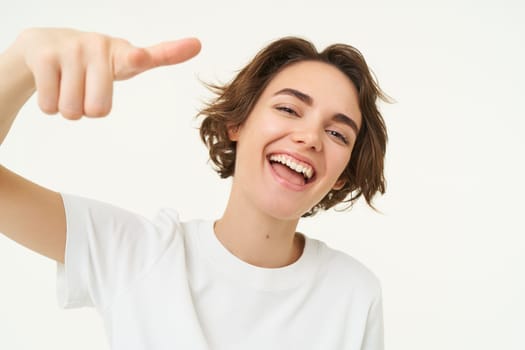 Close up of cheerful, laughing woman, pointing finger at camera and smiling, choosing you, inviting people, standing over white background.