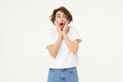 Portrait of screaming girl, holding hands on face, looks shocked and startled by something terrifying, standing over white background. copy space