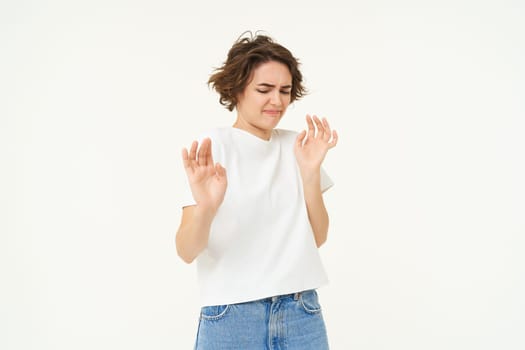 Portrait of woman looking with dislike, rejecting something, raising hands up in defensive gesture, blocking or refusing, standing over white background.