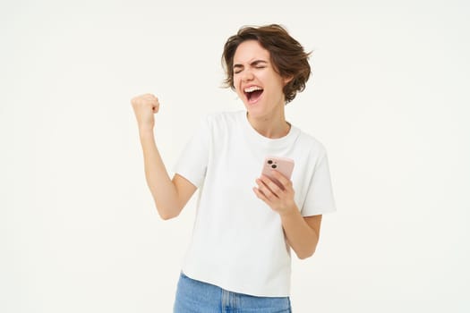Portrait of brunette woman holding mobile phone, winning, triumphing after completing achievement on smartphone app, standing over white background.