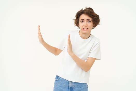 Portrait of awkward, displeased woman shows hand block gesture to the left, avoiding something, refusing, standing over white background.