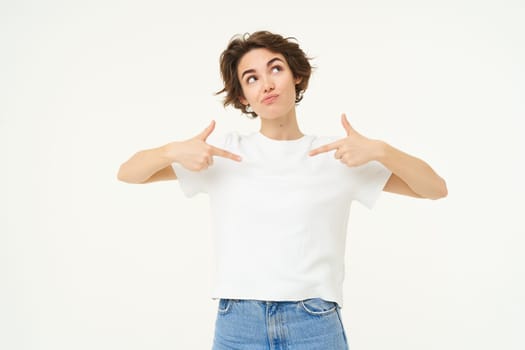 Portrait of thoughtful girl, pointing at herself and looking up, thinking, dreaming, posing over white studio background.