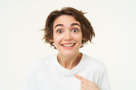 Portrait of girl with surprised face, points at herself amazed, stands isolated over white studio background. Copy space