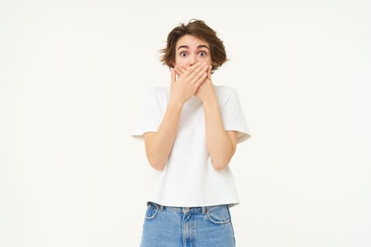 Portrait of woman with surprised face, covers her mouth and gasps shocked, stands isolated over white background.