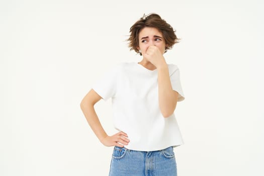 Portrait of woman disgusted by bad smell, shuts her nose, stands over white studio background.