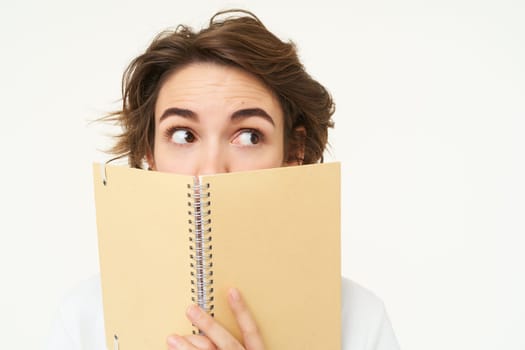Woman of cute brunette woman hiding her face behind planner, peeking aside, looking right at banner, standing over white background. Copy space