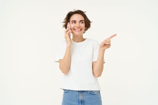 Portrait of friendly, smiling cute girl, pointing finger right, talking on mobile phone, showing smth aside on white copy space, isolated on studio background.