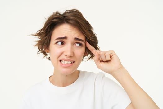 Image of woman cringe, points finger at head, avoids eye contact, thinks someone is crazy, isolated over white background.