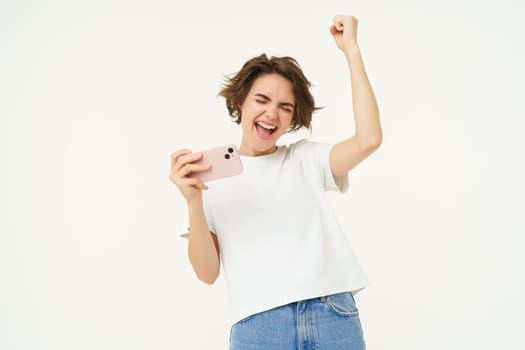 Portrait of excited girl winning mobile phone games and celebrating, laughing and making fist pump, standing over white background. copy space