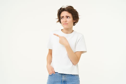 Portrait of woman looking with doubt, pointing left, staring at something with unsure, concerned face expression, standing over white studio background.