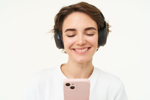 Portrait of happy, smiling beautiful woman, listening to music in wireless headphones, looking at smartphone, choosing song on streaming app, holding mobile phone, standing over white background.