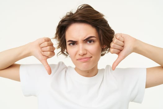 Close up portrait, face of young woman that is showing thumbs down, dislike, do not recommend gesture, express negative feedback, isolated over white background.