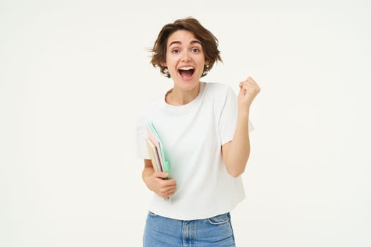 Enthusiastic brunette woman makes fist pump, holds documents and notes, looks thrilled and happy, winning, triumphing, posing over white background.