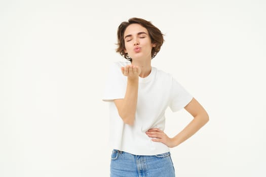 Portrait of young woman sending air kiss, blowing at camera and smiling, standing over white background.