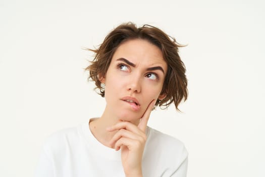 Portrait of thinking woman, touches her chin, looks serious and thoughtful, poses over white background. Copy space