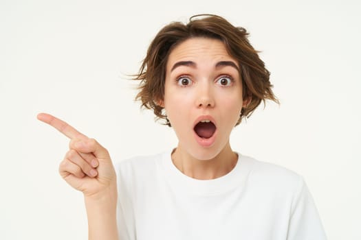 Portrait of woman with surprised face, showing promo to the left, pointing at something, standing over white background. Copy space