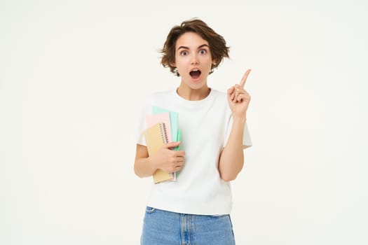 Portrait of woman, student with amazed face, carry her notebooks and documents, points at upper right corner, shows advertisement, studio background.
