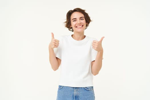 Image of young woman giving positive feedback, shows thumbs up and smiling, looks with confidence, stands over white background. Copy space