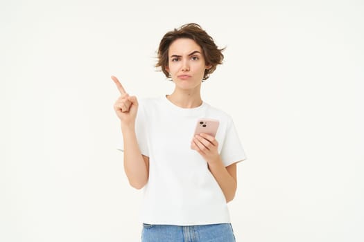 Portrait of woman with smartphone, looking puzzled and pouting, pointing left at banner, thinking, making decision, standing over white background.