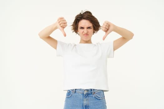 Portrait of disappointed young woman, female client shows dislike gesture, thumbs down, stands over white background.