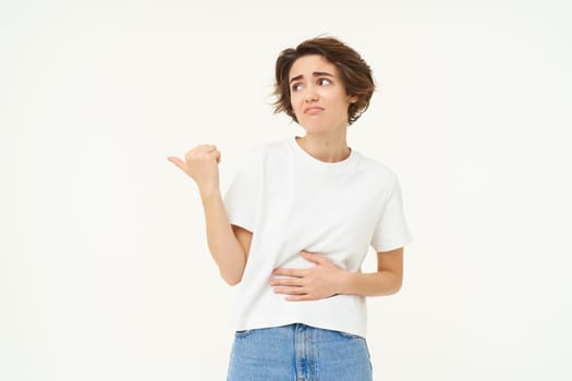 Image of sad woman with stomach ache, points left, shows banner with advertisement, touches her belly, has painful menstrual cramps, isolated over white background.