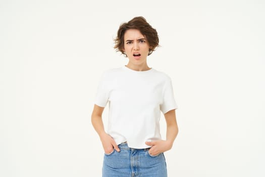 Portrait of arrogant woman, frowning and looking puzzled and disappointed, complaining, holding hands in pants, standing over white studio background.