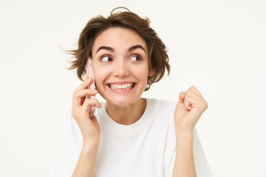 Image of cute brunette woman answer phone call, talking on smartphone, holding mobile near eat and smiling , standing over white background.