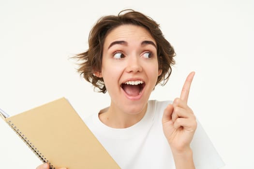 Portrait of happy young woman with planner, student with homework notes, pointing at upper right corner, showing banner and making amazed face expression, standing over white background.