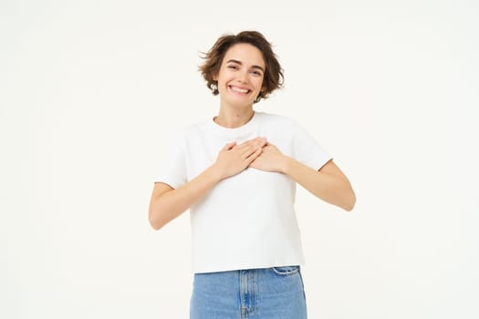 Portrait of woman expressing care, self-love and caring emotions, holds hands on chest, looking with smile at camera, isolated over white background.