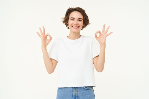 Portrait of young woman, showing okay, ok gestures and smiling, giving positive feedback, recommending, standing over white background.