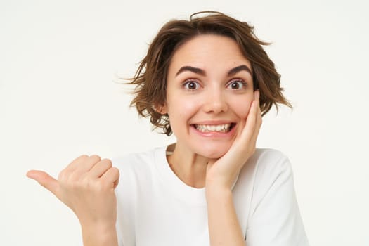 Image of excited brunette woman with short haircut, pointing left, smiling and looking happy, showing you advertisement, standing over white background.