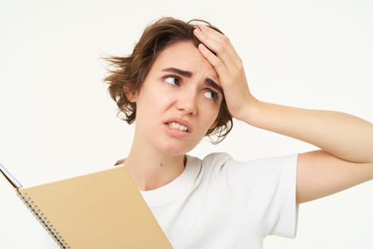 Portrait of puzzled, upset young woman tired of doing homework, holding planner with complicated face, standing over white background.