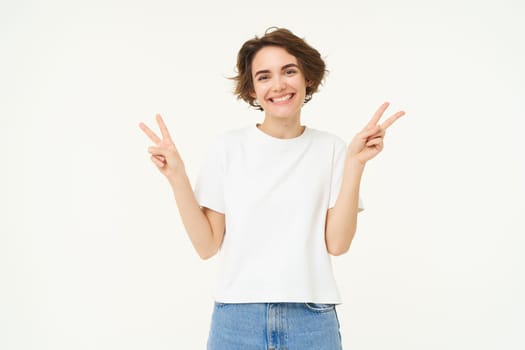 Portrait of happy and carefree woman, shows peace, v-sign gesture, having fun, posing for photo over white background.