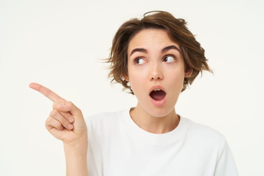 Woman looks surprised, points at left side copy space, shows big sale promo, poses over white studio background.